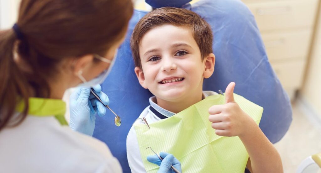 Young boy reclined in a dental chair with dental bib on, giving the camera a big smile and thumbs-up. The image also presents a masked dentist in the foreground, holding a mirror and dental tool, preparing to work on the young boy's teeth.