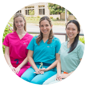 Three dentists sitting outside in brightly-colored scrubs, smiling at a camera.