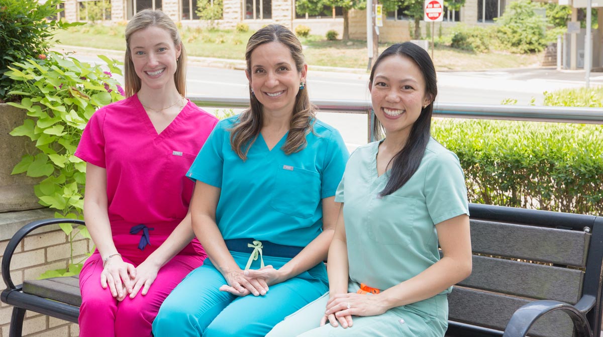 Three dentists sitting outside in brightly-colored scrubs, smiling at a camera.