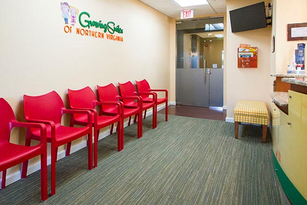 Patient waiting room for Growing Smiles of Northern Virginia with red chairs to the left, a TV hung on the wall, as well as a check in desk for patients on the right.