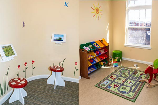 Children's waiting area room. Image is split in half, on the left there are red and white mushroom stools with iPads presented on a wall that has flower stickers. To the right, there is a room full of toys a child would play with, including a toy horse, railroad track map, and bins organized with more toys.