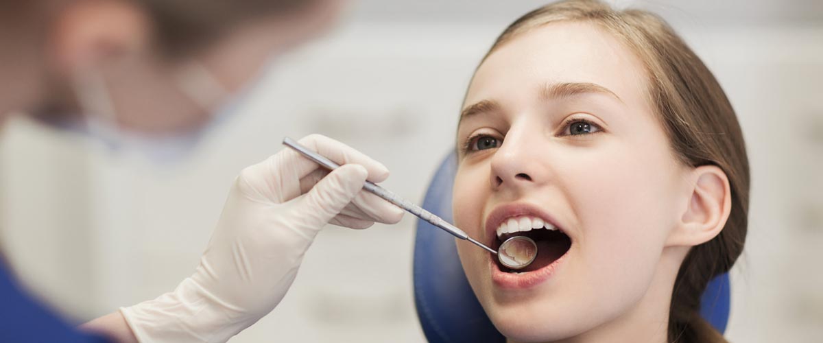 Young girl reclined in a dental chair with her mouth open, as a small mirror is being placed inside her mouth by a gloved-dentist to examine her teeth.