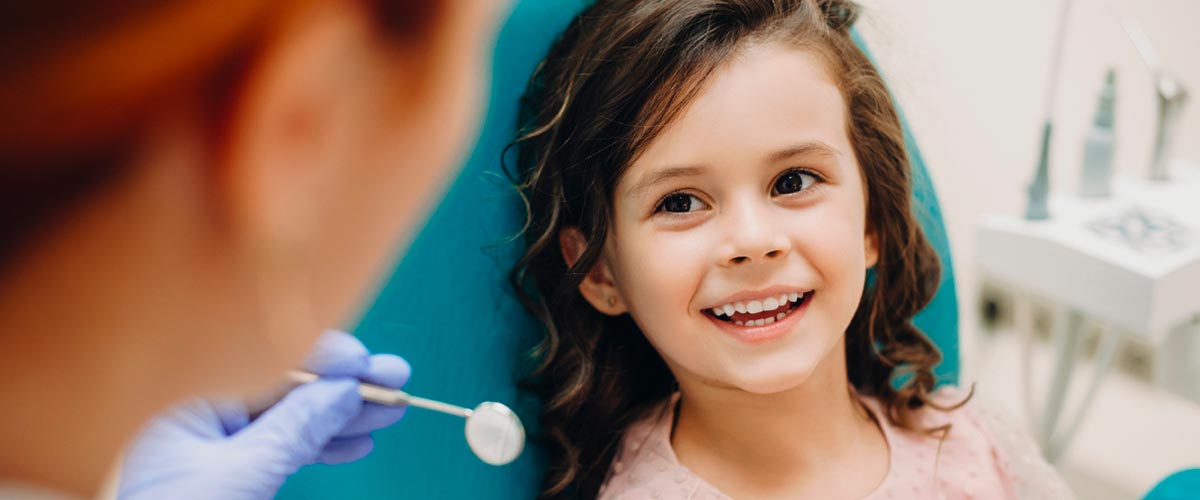 Young girl with curly brown hair and wide eyes, looking up and smiling at the dentist who is holding a mirror to look into her mouth.