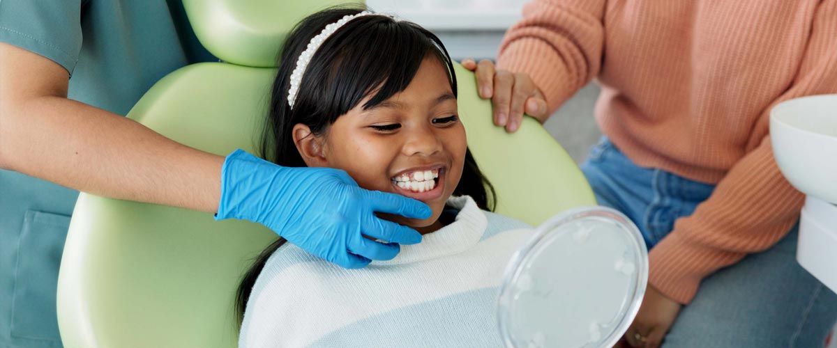 Young girl sitting in a dentist chair, gleaming, as she smiles into a mirror she is holding while a dentist is showing her aspects of her smile.